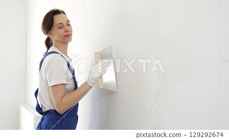 Woman construction worker wearing protective gloves and blue construction coveralls, spreading plaster smoothly across wall using professional drywall taping knife during renovation project. Portrait Woman construction worker wearing protective gloves and blue construction coveralls, spreading plaster smoothly across wall using professional drywall taping knife during renovation project. Portrait 129292674