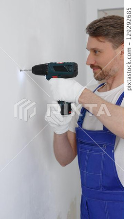 Man construction worker in a blue jumpsuit, safety goggles, and work gloves is screwing a screw into the wall using a cordless screwdriver, carrying out a careful home renovation. Vertical portrait 129292685