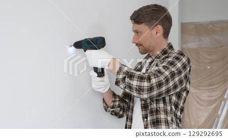 Male construction worker in checked shirt and safety gloves is screwing a screw into the wall using a cordless screwdriver, carrying out a careful home renovation. Portrait view 129292695