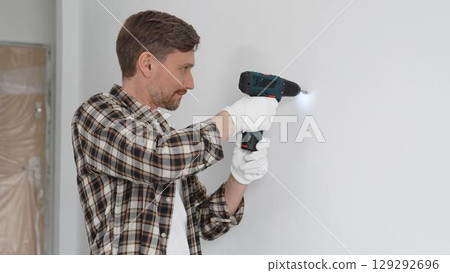 Man construction worker in checked shirt and safety gloves is screwing a screw into the wall using a cordless screwdriver, carrying out a careful home renovation. Horizontal portrait view 129292696
