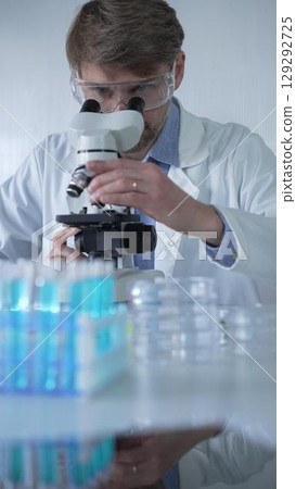 Male scientist researcher wearing lab coat and protective glasses, is using microscope in modern laboratory setup, portrait view. Low key lighting photo. Science concept Male scientist researcher wearing lab coat and protective glasses, is using microscope in modern laboratory setup, portrait view. Low key lighting photo. Science concept 129292725