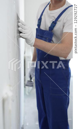 Close-up of man construction worker wearing gloves and blue overalls is using a putty knife to apply plaster on a white wall, performing home renovation and improvement 129292727