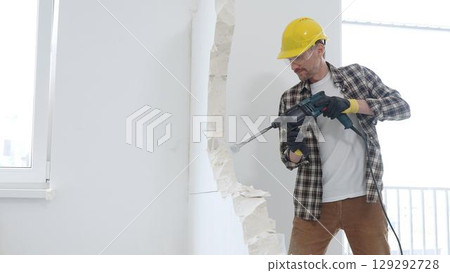 Male construction worker wearing beige checkered shirt, yellow hard hat and protective gloves, is demolishing white wall with rotary hammer drill, generating dust, medium long shot. Renovation project 129292728