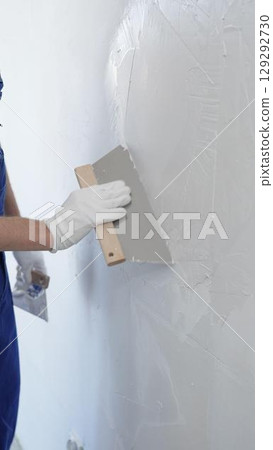 Close up of the construction worker hand wearing protective gloves, spreading plaster smoothly across wall using professional drywall taping knife realizing renovation project, vertical view 129292730