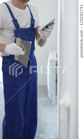 Man construction worker wearing protective gloves and blue construction coveralls, spreading plaster smoothly across wall using professional drywall taping knife Man construction worker wearing protective gloves and blue construction coveralls, spreading plaster smoothly across wall using professional drywall taping knife 129292731