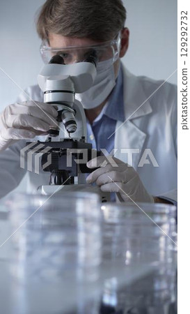 Man scientist wearing lab coat, white gloves, medical mask and protective glasses using microscope doing research in modern laboratory, portrait view. Low key lighting photo. Science and Medicine 129292732