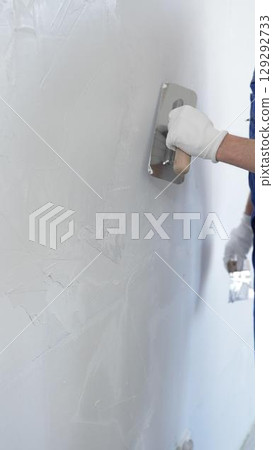 Professional construction worker wearing blue work overall is using a finishing trowel to carefully apply plaster on a wall, demonstrating expertise in home renovation 129292733