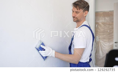 Man construction worker wearing protective white gloves and blue construction coveralls, is sanding white wall with blue hand block, preparing smooth surface during interior home renovation project 129292735