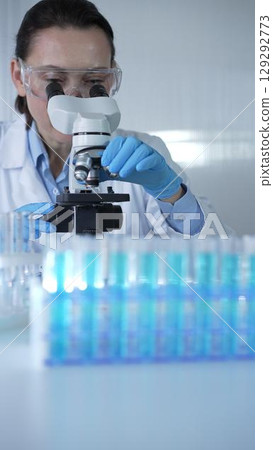 Female scientist wearing lab coat and protective blue gloves using microscope doing research in modern laboratory, portrait view. Low key lighting photo. Science and Medicine Female scientist wearing lab coat and protective blue gloves using microscope doing research in modern laboratory, portrait view. Low key lighting photo. Science and Medicine 129292773