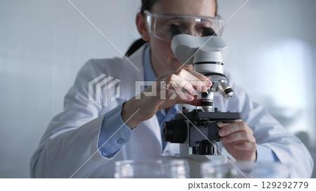 Woman scientist wearing lab coat and protective glasses is using microscope in modern laboratory setup. Low key lighting photo. Science and Medicine Woman scientist wearing lab coat and protective glasses is using microscope in modern laboratory setup. Low key lighting photo. Science and Medicine 129292779