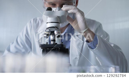 Man scientist researcher wearing white gloves, protective glasses and medical mask is using microscope in modern laboratory, portrait view. Low key lighting photo. Science and Medicine 129292784