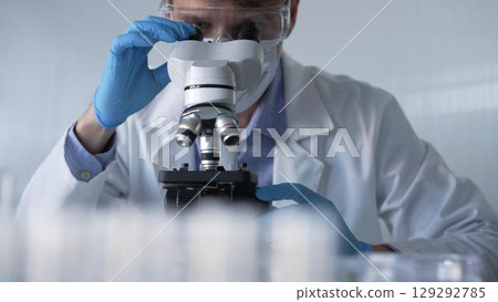 Male scientist researcher wearing blue gloves, protective glasses and medical mask, is using microscope in modern laboratory, portrait view. Low key lighting photo. Science and Medicine 129292785