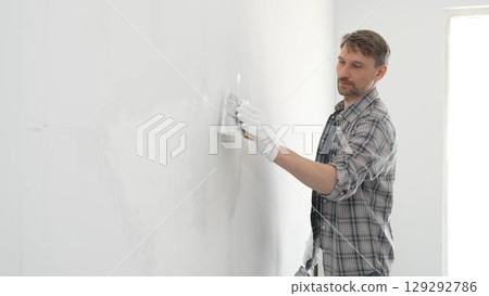 Construction worker spreading plaster on white wall, wearing protective gloves and t-shirt during home renovation project, working with professional skill and focus Construction worker spreading plaster on white wall, wearing protective gloves and t-shirt during home renovation project, working with professional skill and focus 129292786