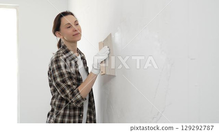 Female construction professional wearing checkered shirt is smoothing white wall surface, applying putty with professional spatula technique, showcasing skilled interior renovation work 129292787