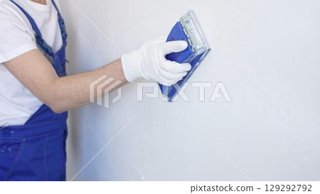 Unknown male construction worker wearing protective white gloves and blue construction coveralls, is sanding white wall with blue hand block, preparing smooth surface during interior home renovation 129292792