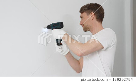 Professional male worker wearing white t-shirt and protective gloves, is drilling hole in a wall with cordless drill during home renovation work. Horizontal portrait view Professional male worker wearing white t-shirt and protective gloves, is drilling hole in a wall with cordless drill during home renovation work. Horizontal portrait view 129292794