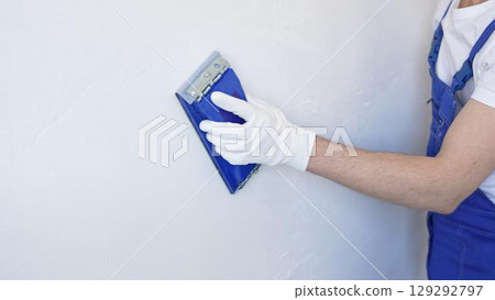 Unknown male construction worker wearing protective white gloves and blue construction coveralls, is sanding white wall with blue hand block, preparing smooth surface during interior home renovation 129292797