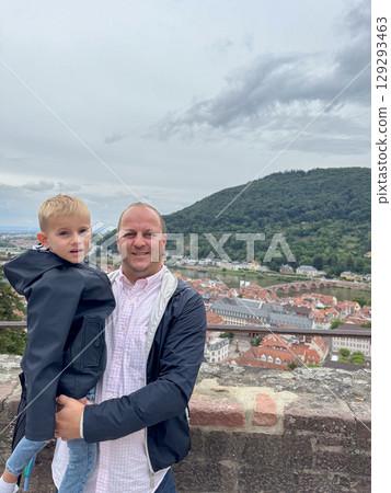 Father holds his young son as they stand on a terrace overlooking a picturesque cityscape. The pair enjoy quality time together while embracing a beautiful mountainous backdrop Father holds his young son as they stand on a terrace overlooking a picturesque cityscape. The pair enjoy quality time together while embracing a beautiful mountainous backdrop 129293463