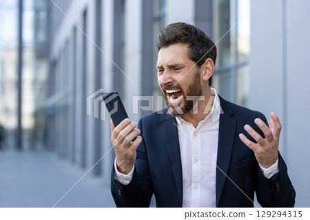 A distressed businessman in a suit vents frustration at his smartphone outside modern offices during a workday. Captures a moment of urban stress and communication breakdown. A distressed businessman in a suit vents frustration at his smartphone outside modern offices during a workday. Captures a moment of urban stress and communication breakdown. 129294315