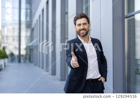 Confident male professional extending a handshake outdoors in a corporate office setting, showcasing business friendliness and a welcoming attitude. Confident male professional extending a handshake outdoors in a corporate office setting, showcasing business friendliness and a welcoming attitude. 129294316