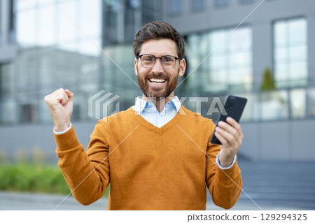 Happy man in casual attire rejoices while holding a smartphone outdoors near office buildings. Happy man in casual attire rejoices while holding a smartphone outdoors near office buildings. 129294325