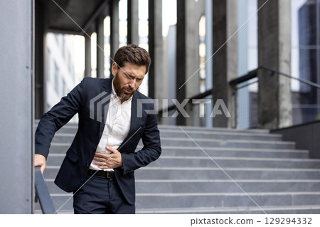 A professional businessman in formal wear gripping his stomach in visible discomfort, standing outdoors near urban office stairs, expressing feelings of pain, unease, and potential health concerns. 129294332