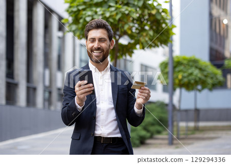 Smiling man in professional attire using a smartphone and credit card, representing online shopping or digital transactions, standing in a modern urban environment with a bright and sunny ambience. 129294336