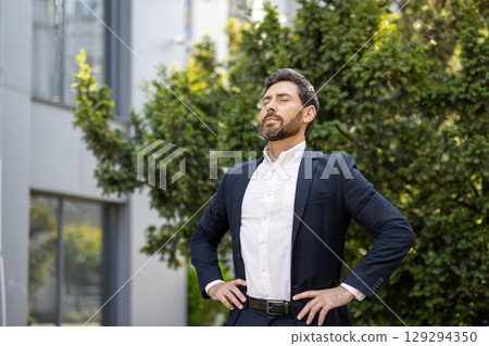 A man in formal attire takes a moment outdoors, with hands on hips, to enjoy the fresh air, exuding confidence and a peaceful demeanor, backed by urban greenery near an office. 129294350