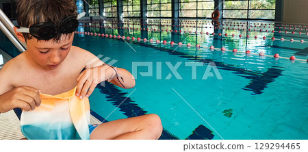 A playful boy holds a torn swimming cap from the pool in his hands. Concept of swimming equipment, children's sports, a joyful moment from childhood associated with sports. A playful boy holds a torn swimming cap from the pool in his hands. Concept of swimming equipment, children's sports, a joyful moment from childhood associated with sports. 129294465