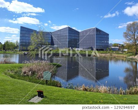 Modern office buildings with angular designs reflect in a calm pond. A grassy lawn with an orange and black charging station for a lawnmower robot 129294697