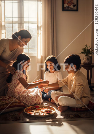 A mother, in her 30s, lovingly watches as her two young daughters, smiling with joy, tie a thread bracelet on their brother's wrist 129295448