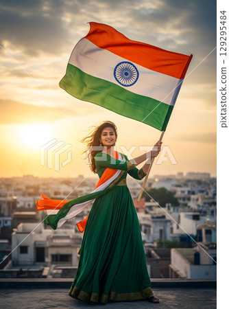 A happy young Indian woman holding a flag aloft at sunset, standing on a rooftop in a beautiful pose 129295484