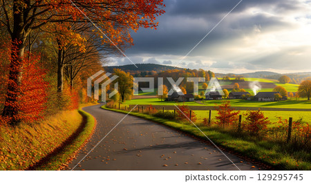 A winding road passes through a picturesque rural landscape with small houses and rolling green hills, surrounded by vibrant autumn trees and sunlit fields 129295745