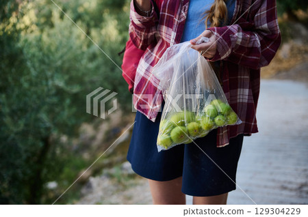 Person Holding Fresh Green Fruit in Plastic Bag Outdoors Surrounded by Nature 129304229