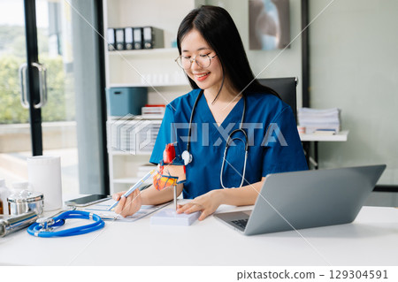 Confident young female doctor in white medical uniform sit at desk working on computer. Smiling use laptop Confident young female doctor in white medical uniform sit at desk working on computer. Smiling use laptop 129304591