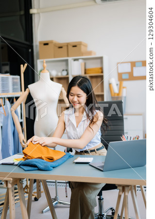 Asian young woman on desk in office of fashion designer and holds tablet, laptop and smartphone Asian young woman on desk in office of fashion designer and holds tablet, laptop and smartphone 129304604