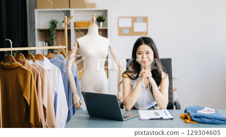 fashion designer. Attractive woman with smile while standing in workshop. Beautiful owner woman working and holds tablet, laptop and smartphone in studio. fashion designer. Attractive woman with smile while standing in workshop. Beautiful owner woman working and holds tablet, laptop and smartphone in studio. 129304605