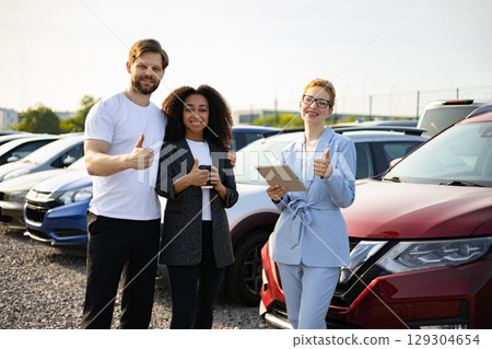 Three people at a car dealership giving a thumbs up in front of a red car, showcasing customer satisfaction. 129304654