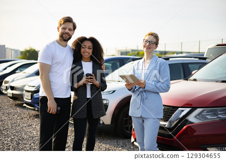A car salesperson assists a couple in a car lot. The three are smiling as they look at the camera. 129304655