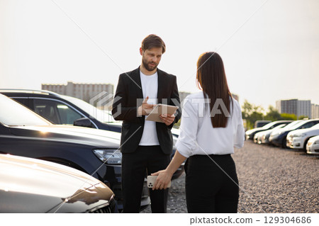 A male car dealer, dressed in a suit, is showing a tablet to a female customer at a car dealership. They are surrounded by various cars in the parking lot. 129304686