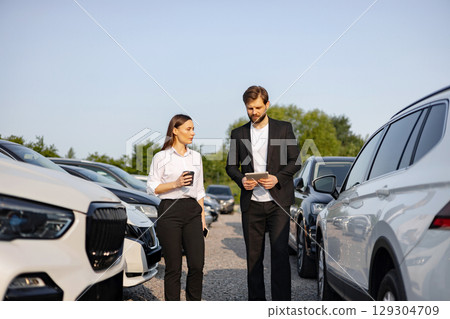 A male car dealer shows a vehicle to a woman on a sunny day at a car dealership. The woman is holding a coffee cup. 129304709