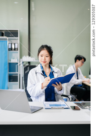 Confident young asian female doctor in white medical uniform sit at desk working on computer. 129305087