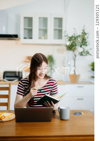 Asian woman working remotely on a laptop and tablet in a cozy kitchen workspace. Perfect for education Asian woman working remotely on a laptop and tablet in a cozy kitchen workspace. Perfect for education 129305125