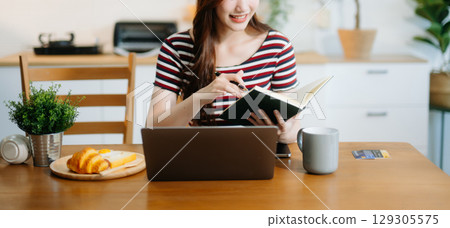 Asian woman working remotely on a laptop and tablet in a cozy kitchen workspace. Perfect for education Asian woman working remotely on a laptop and tablet in a cozy kitchen workspace. Perfect for education 129305575