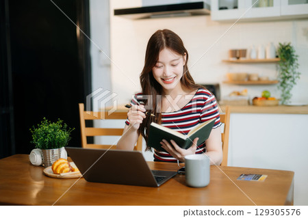 Asian woman working remotely on a laptop and tablet in a cozy kitchen workspace. Perfect for education Asian woman working remotely on a laptop and tablet in a cozy kitchen workspace. Perfect for education 129305576