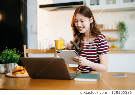 Happy woman with headphones working on a tablet in a bright kitchen. Ideal for themes of remote work Happy woman with headphones working on a tablet in a bright kitchen. Ideal for themes of remote work 129305586