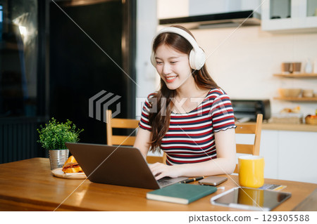 Asian woman working remotely on a laptop and tablet in a cozy kitchen workspace. Perfect for education Asian woman working remotely on a laptop and tablet in a cozy kitchen workspace. Perfect for education 129305588