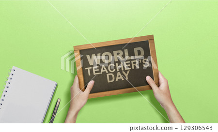 Human hand holding a chalkboard with the message of World Teacher Day on a green background. Concept of World Teacher Day Human hand holding a chalkboard with the message of World Teacher Day on a green background. Concept of World Teacher Day 129306543