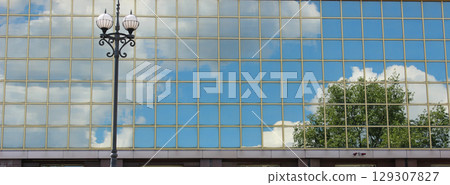 Reflection Of Trees, Sky And Clouds In Glass Facade Of Modern Building With Street Lamp In Front  129307827