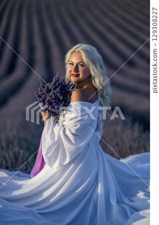 Blonde woman poses in lavender field at sunset. Happy woman in white dress holds lavender bouquet. Aromatherapy concept, lavender oil, photo session in lavender 129308227